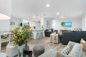 A modern living room with a grey sofa, a white dining area, and a kitchen in the background. - Fall Resort Getaway! 542 Augusta Dr. w/ 6 Seat Golf Cart on Golf Course! (Miramar Beach)