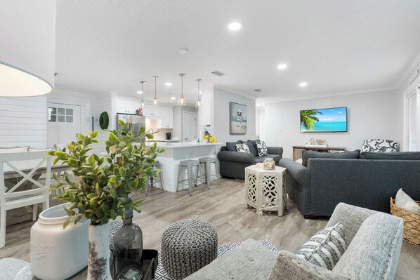 A modern living room with a grey sofa, a white dining area, and a kitchen in the background.