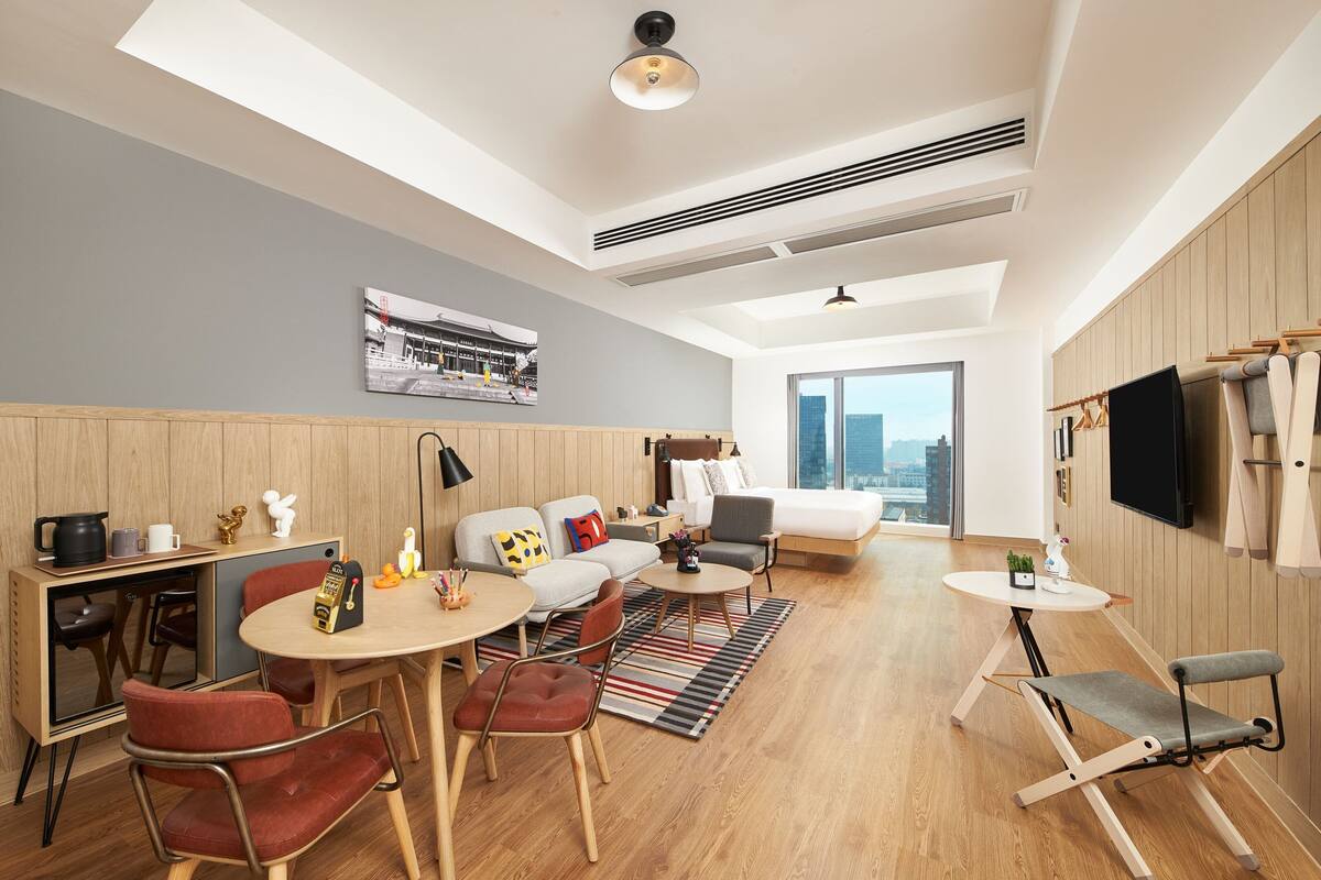 Quarto de hotel moderno com área de refeições, sofá, cama e televisão fixada na parede.