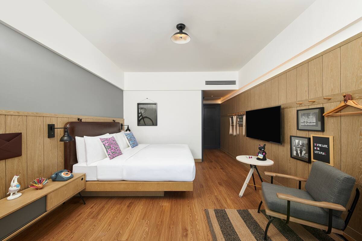 Quarto de hotel moderno com cama de madeira, uma pequena escrivaninha, uma cadeira, televisão e cabideiro fixado na parede.
