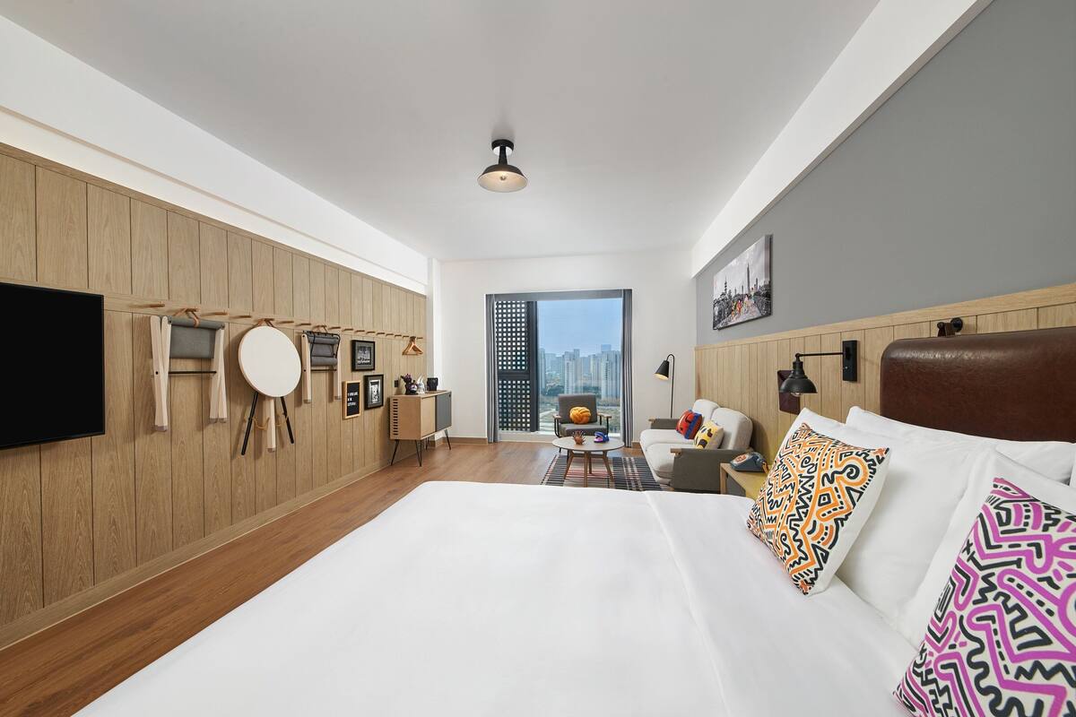 Quarto de hotel moderno com uma cama grande, televisão de tela plana, escrivaninha com cadeira e vista da cidade por uma janela ampla.