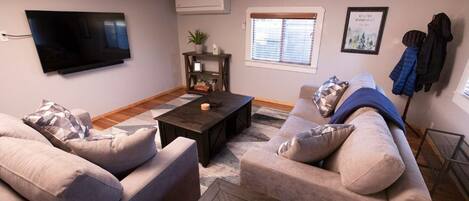 A living room with a flat-screen TV, a dark coffee table, two sofas, a small side table, and a window with blinds.