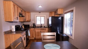 Cocina compacta con armarios de madera, un refrigerador negro, una mesa de comedor con dos sillas y una ventana con persianas.