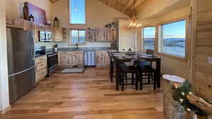 A spacious kitchen with wooden cabinets, a stainless steel refrigerator, a microwave, and a dishwasher. There is a dining area with a table and chairs, and large windows offering views of the outdoors.