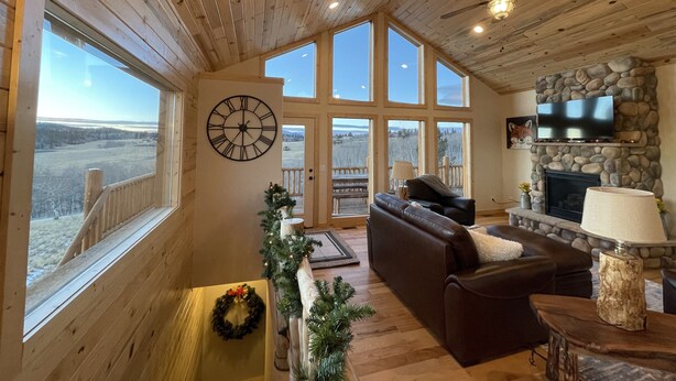 A spacious living room with a large wooden ceiling, a stone fireplace, a television mounted on the wall, and a view of a snowy landscape through large windows.