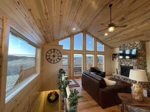 A spacious living room with a large wooden ceiling, a stone fireplace, a television mounted on the wall, and a view of a snowy landscape through large windows.