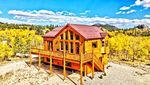 A wooden cabin with a red roof, a porch, and a view of yellow trees and mountains.