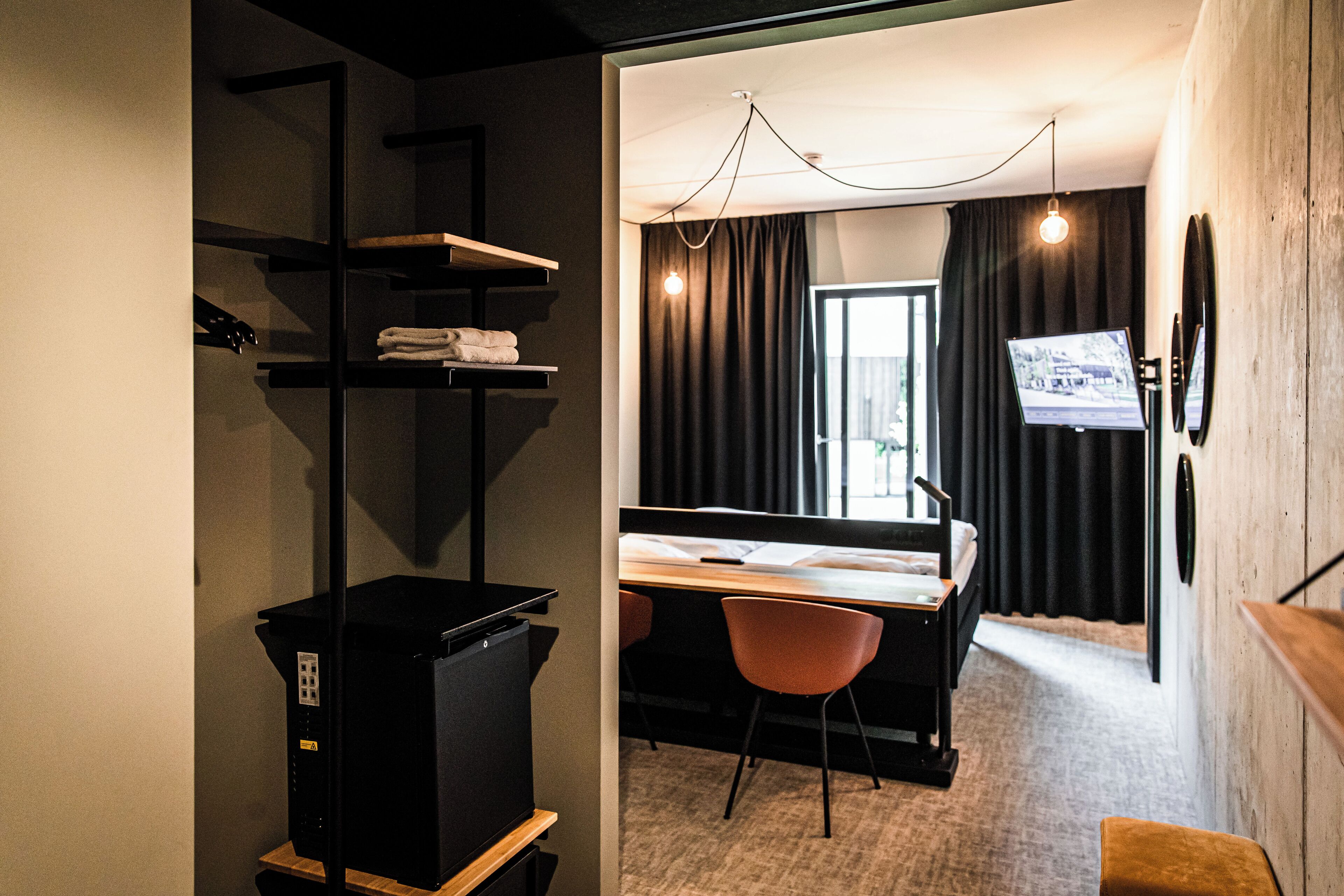 A hotel room with a desk, chair, and a TV mounted on the wall. There is a window with curtains, a shelf with towels, and a trash bin.