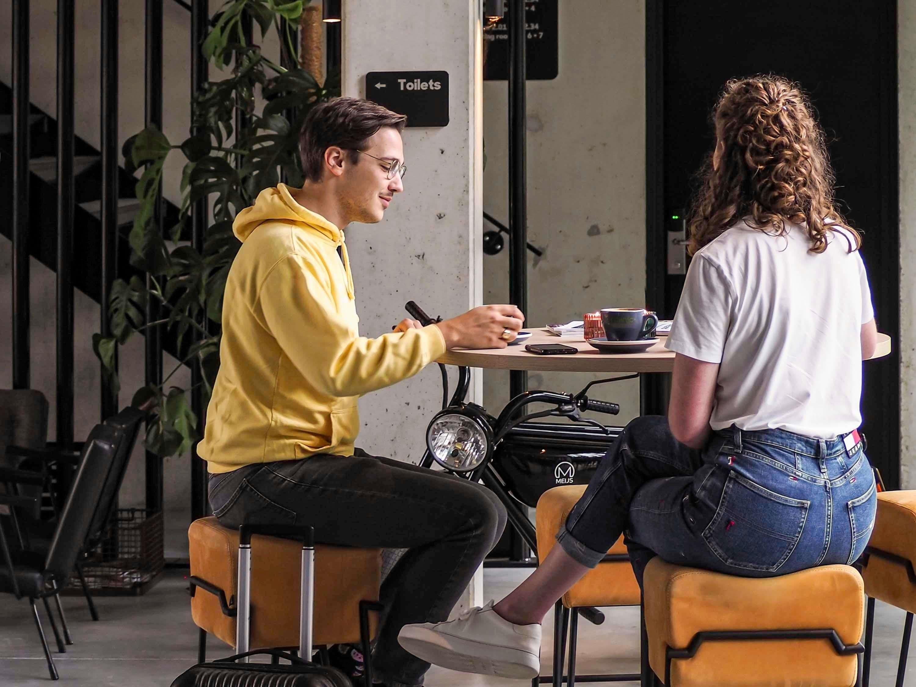 Dos personas sentadas en una mesa con una taza de café, una bicicleta y un letrero que indica los servicios sanitarios al fondo.