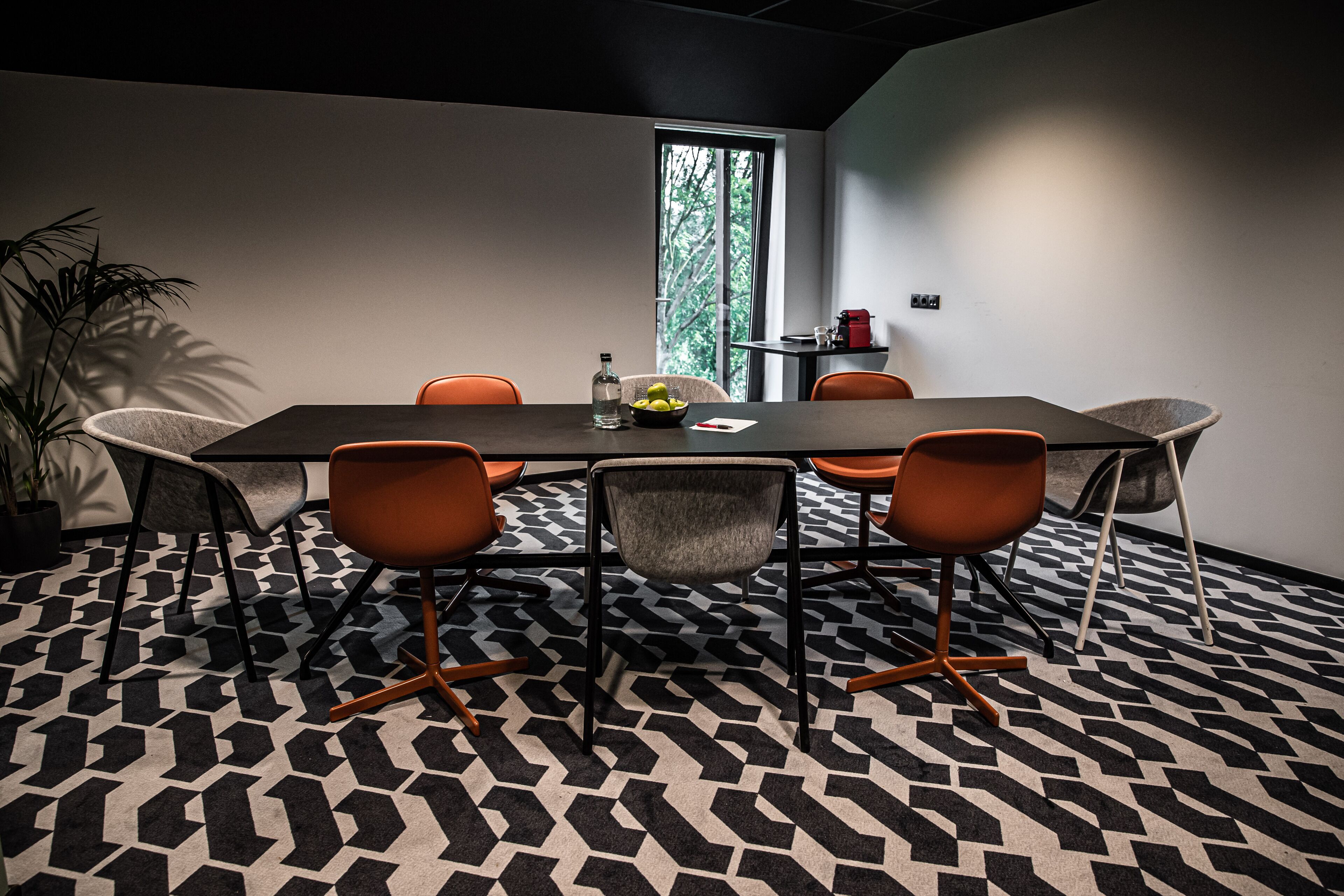 Uma sala de conferências com carpete estampado em preto e branco, uma mesa retangular e quatro cadeiras laranja.