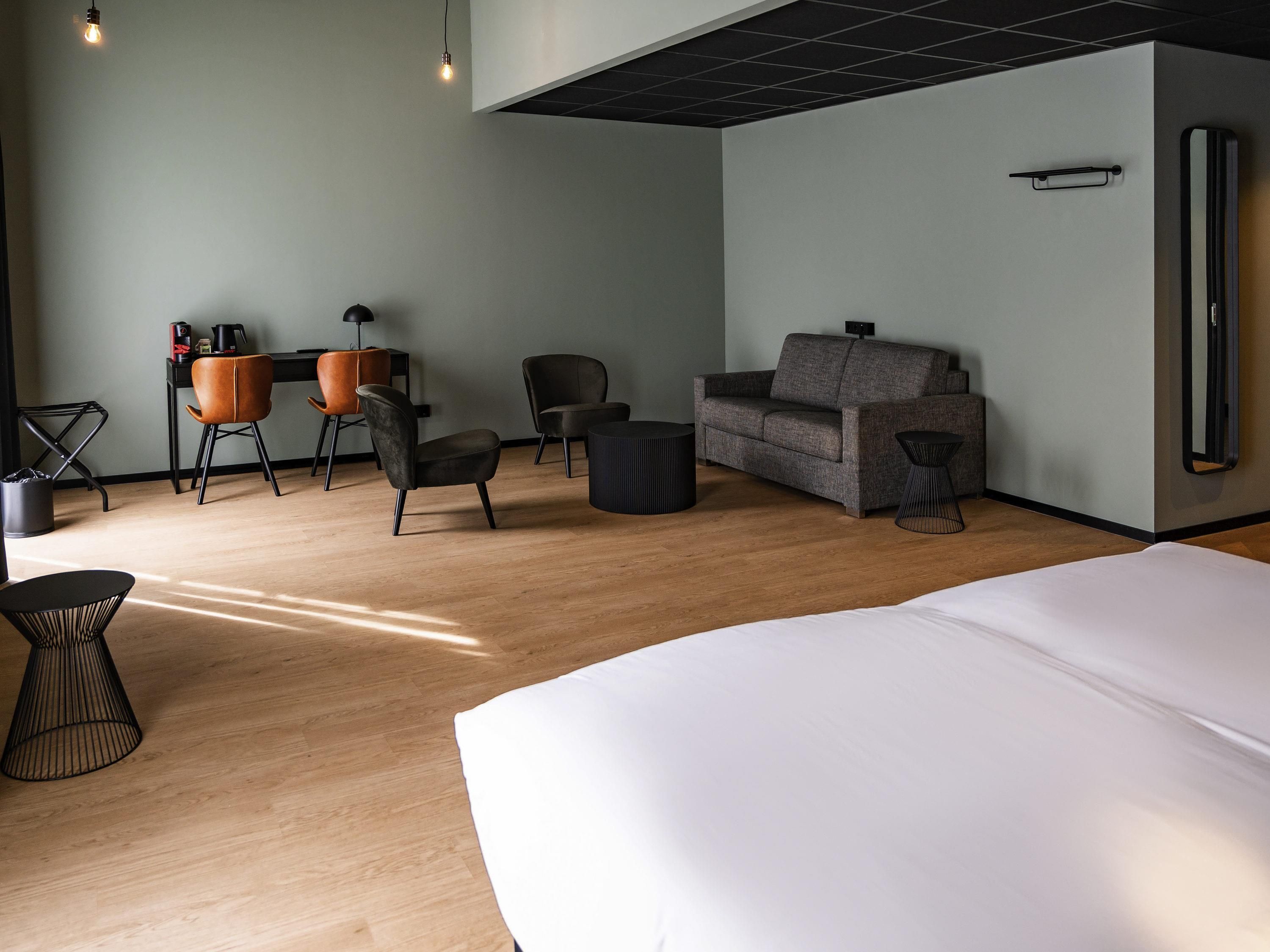 A modern hotel room with a wooden floor, a sofa, a desk with chairs, a bed, and a small table.