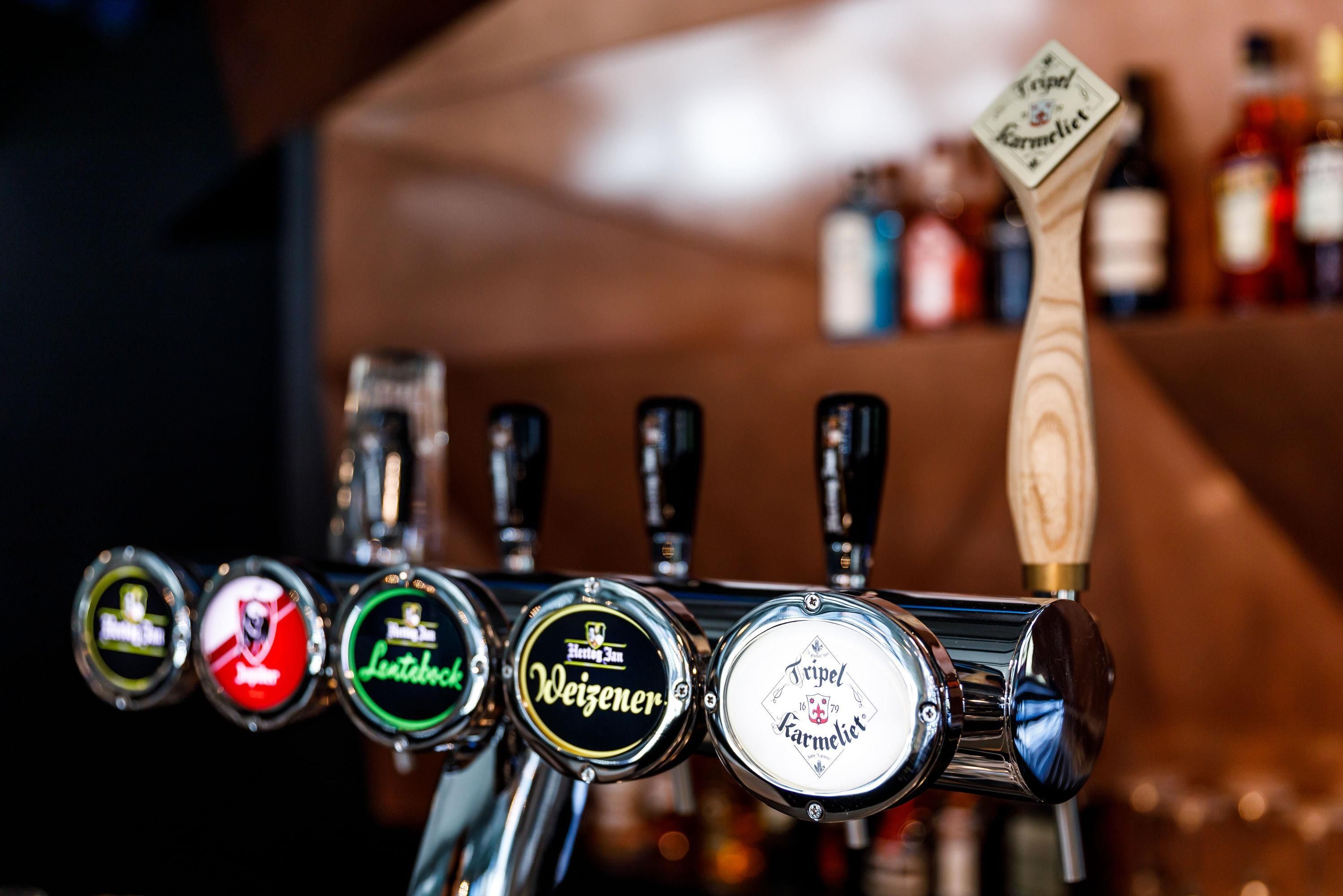 Primer plano de un grifo de cerveza con varias etiquetas, incluyendo 'Weizen' y 'Karmeliet'.