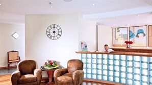 Un área de recepción con un reloj, dos sillones, un escritorio y una pared con obras de arte enmarcadas.