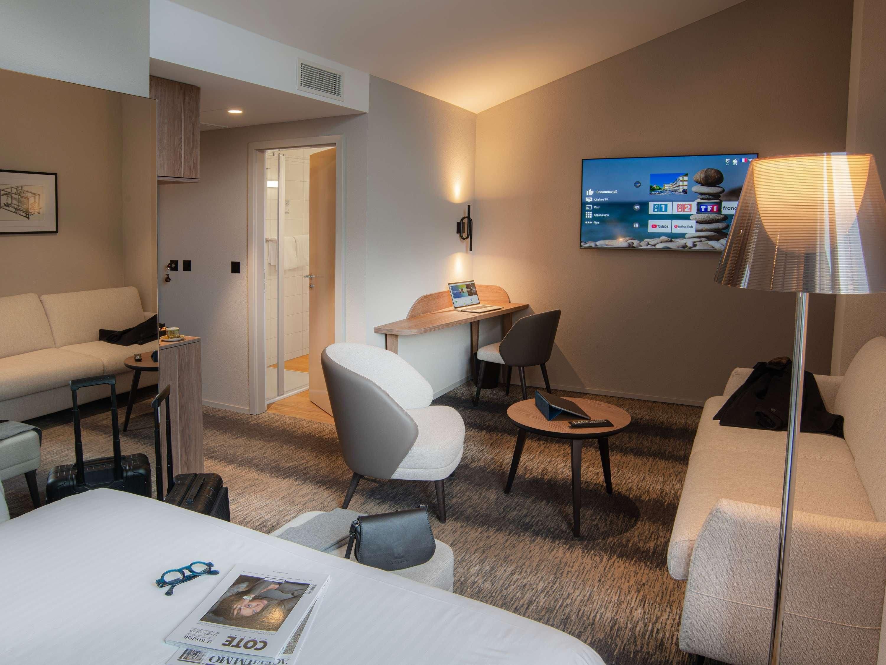 A hotel room with a bed, a desk, a chair, a sofa, a TV, and a lamp.