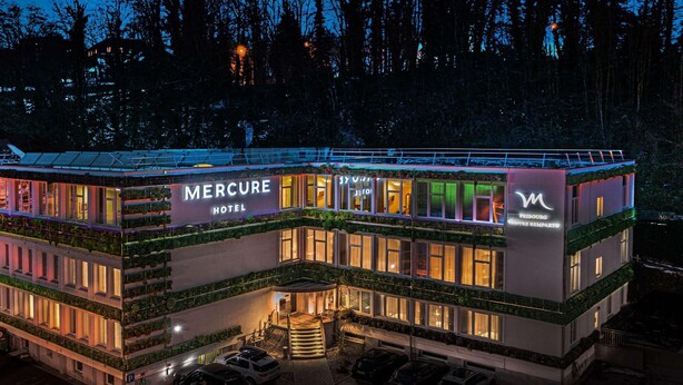 A multi-story hotel with a prominent 'Mercure Hotel' sign, a parking area, and a building with a green facade. - Mercure Fribourg Centre Remparts (Fribourg)