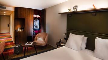 A hotel room with a large bed, a small armchair, a desk with a lamp, a wardrobe, and a colorful carpet.