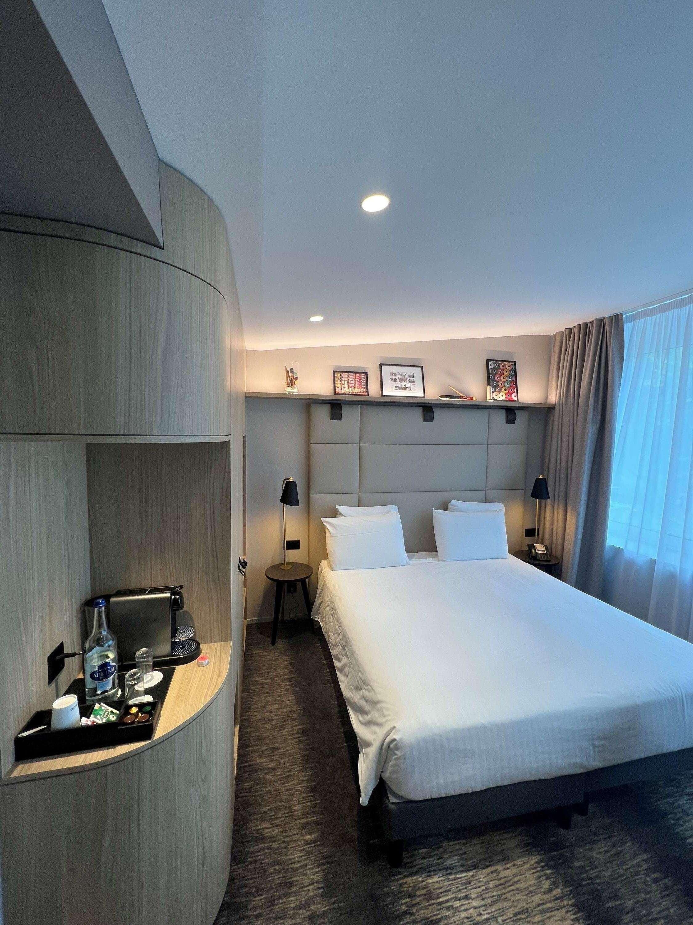 A hotel room with a bed, bedside tables, a desk with a coffee machine, and a wall-mounted shelf.