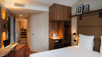 A hotel room with a bed, desk, laptop, chair, wardrobe, and a hallway leading to another room.