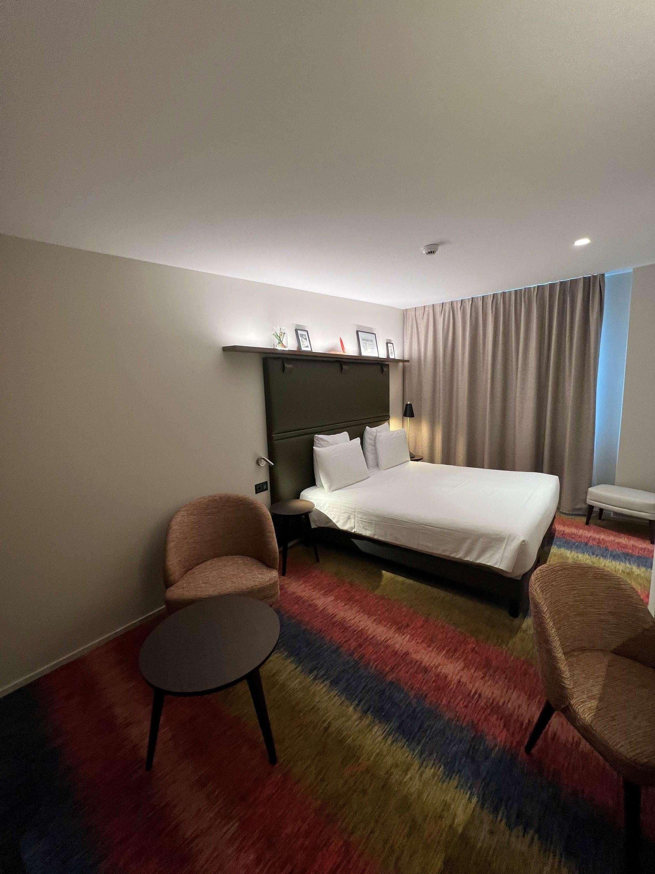 A hotel room with a bed, two armchairs, a small table, a headboard with pictures, and a large window with curtains.