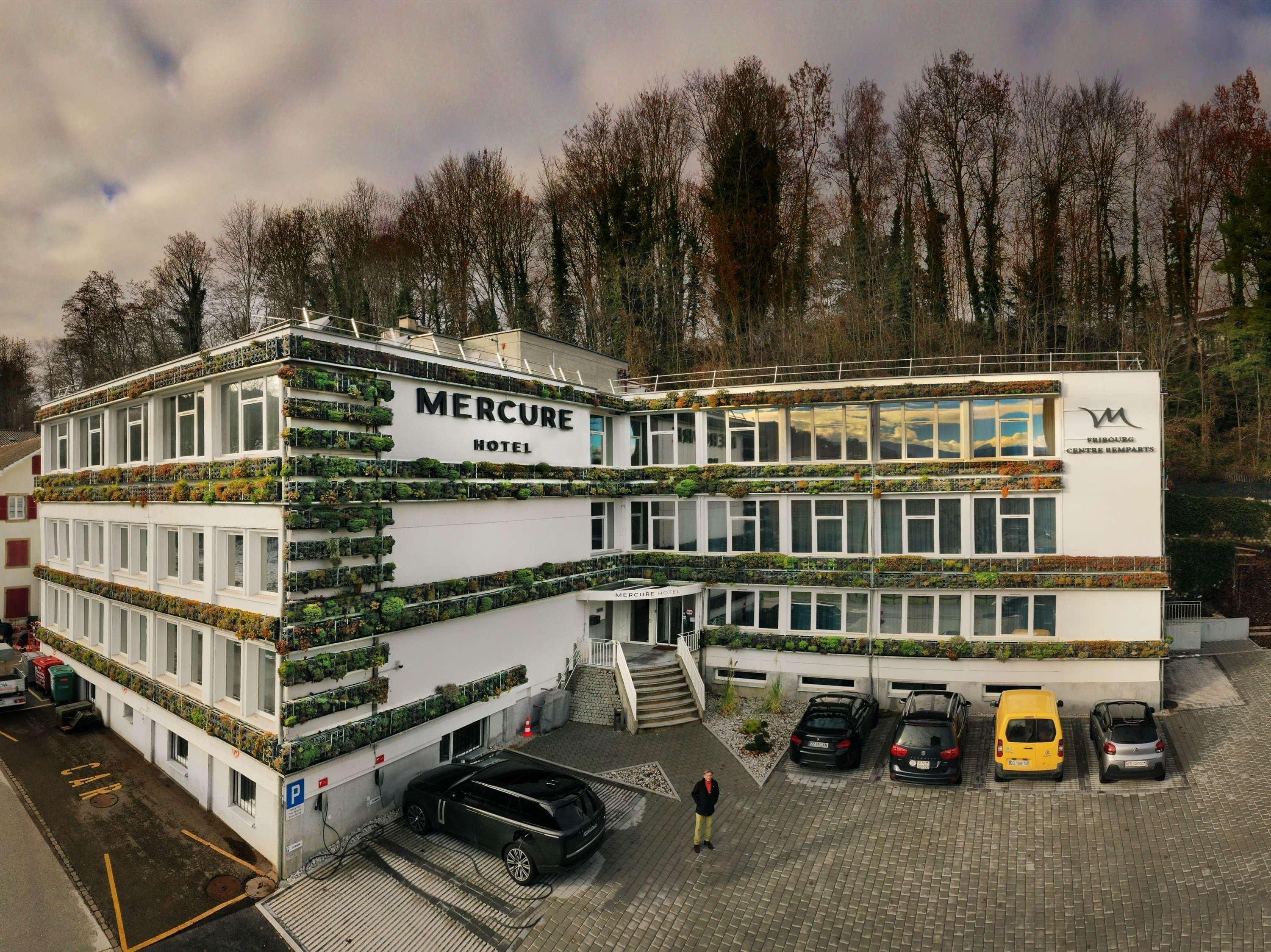 A multi-story hotel with a white facade, balconies with greenery, and a parking area with several cars.