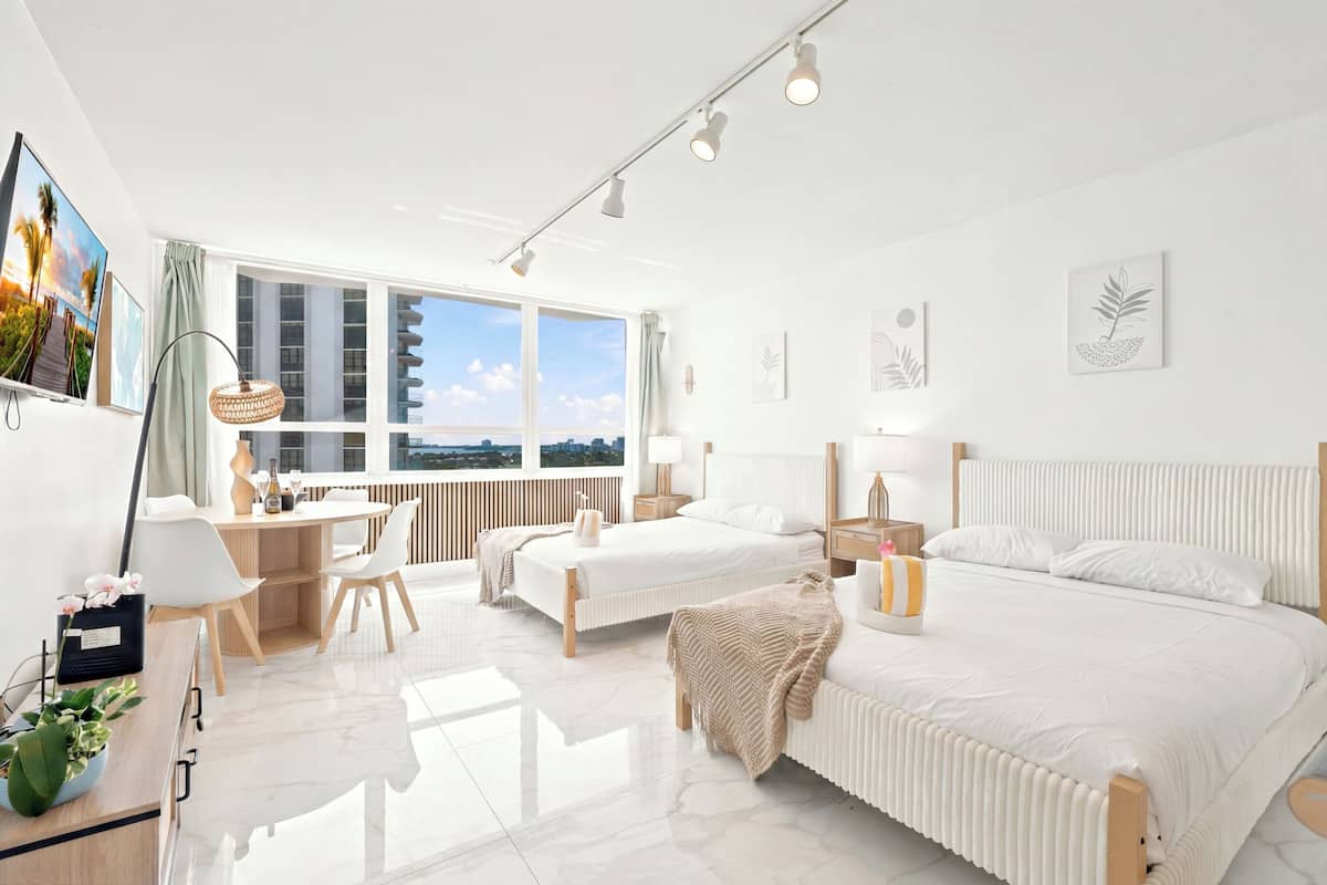 A modern hotel room with a large window, a desk, two beds, and a view of the cityscape.