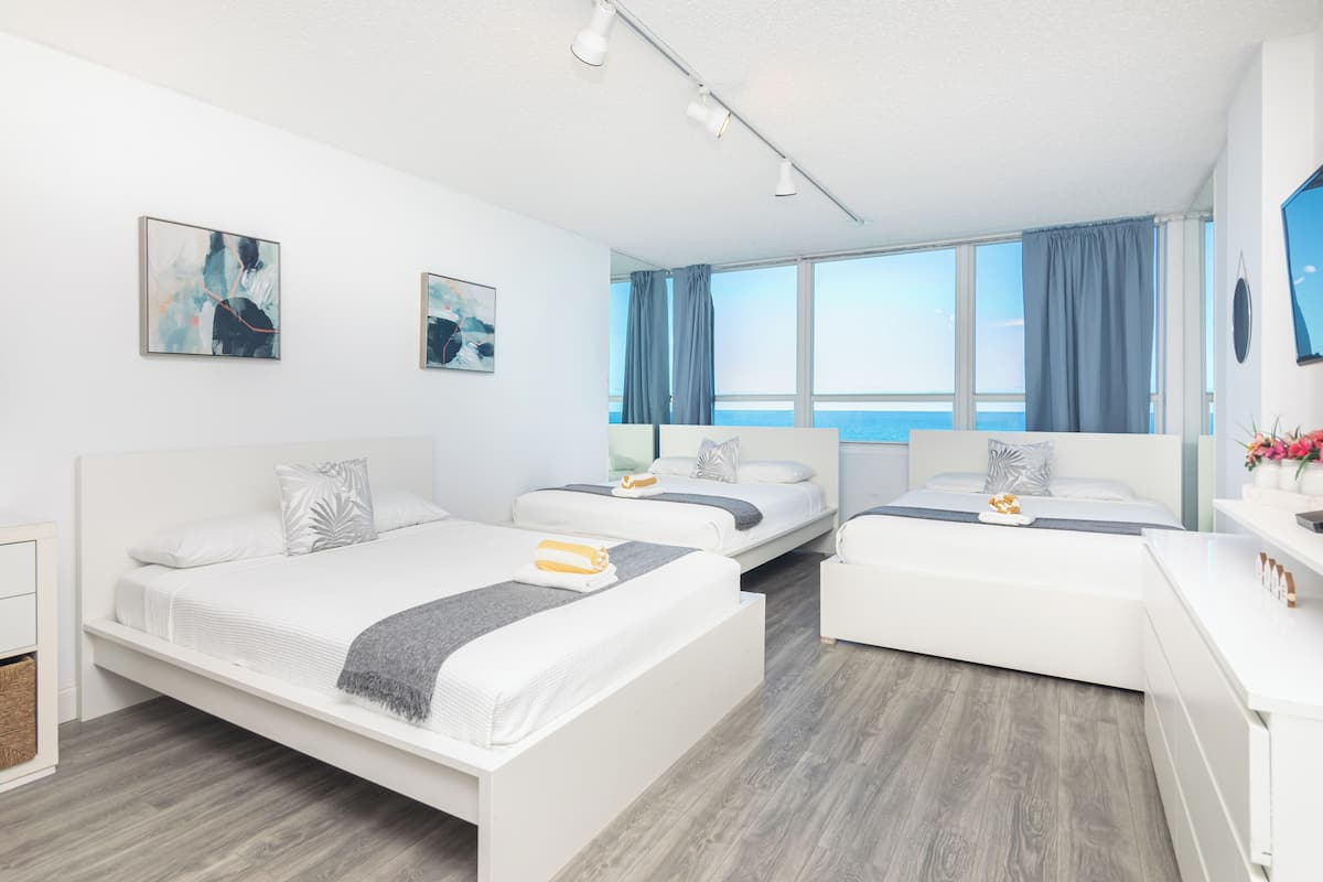A modern hotel room with two beds, a desk, and a large window offering a view of the sea.