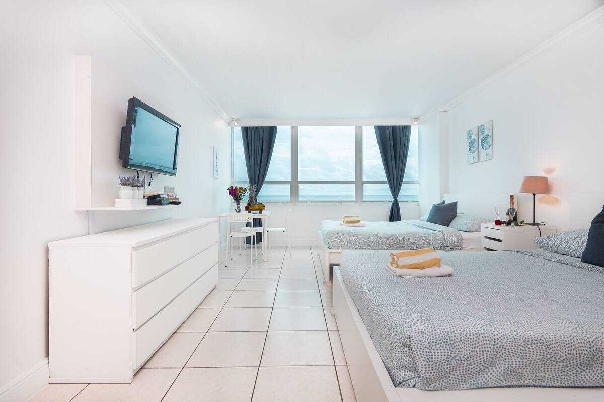 A modern bedroom with a bed, bedside tables, a TV, and a view of the sea.