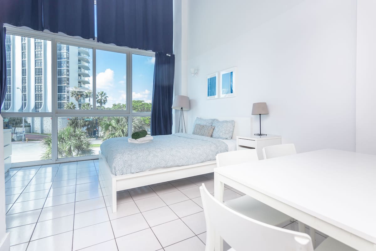 A modern bedroom with a large window, a bed, a bedside table, and a dining area.