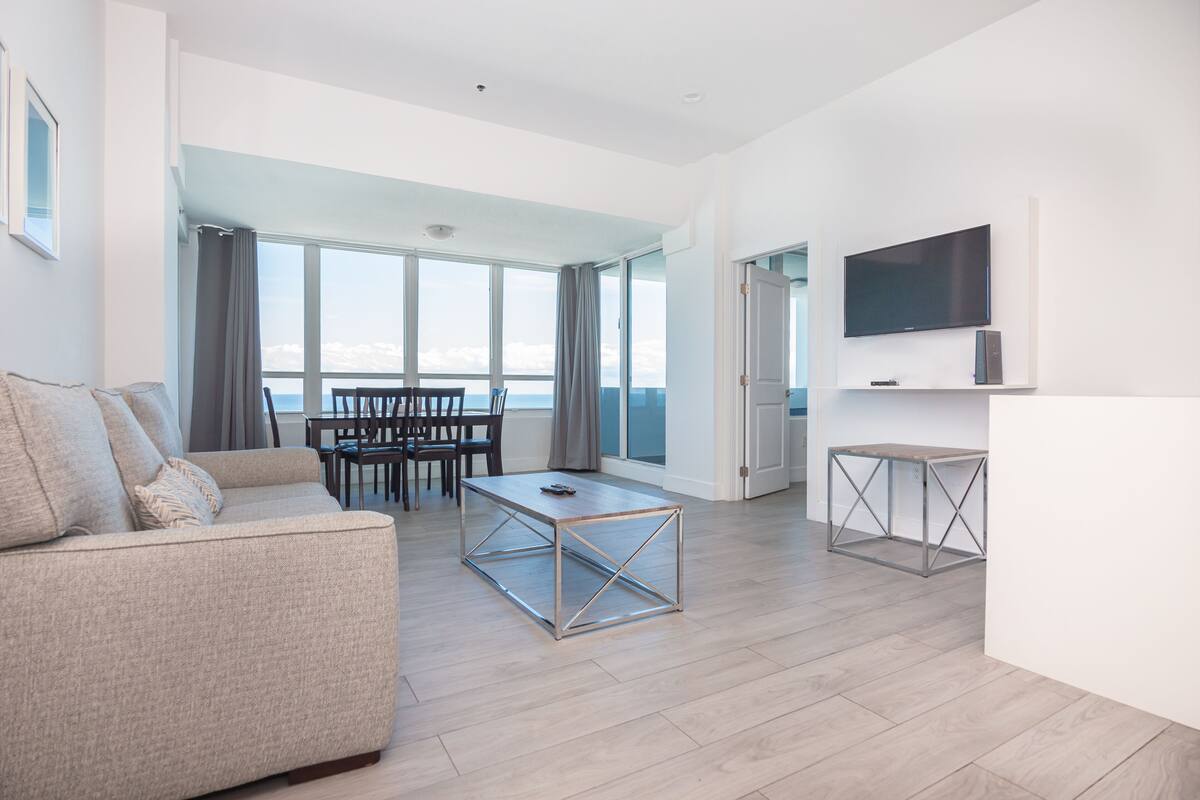 A modern living room with a sofa, coffee table, and TV. There is a balcony with a view.