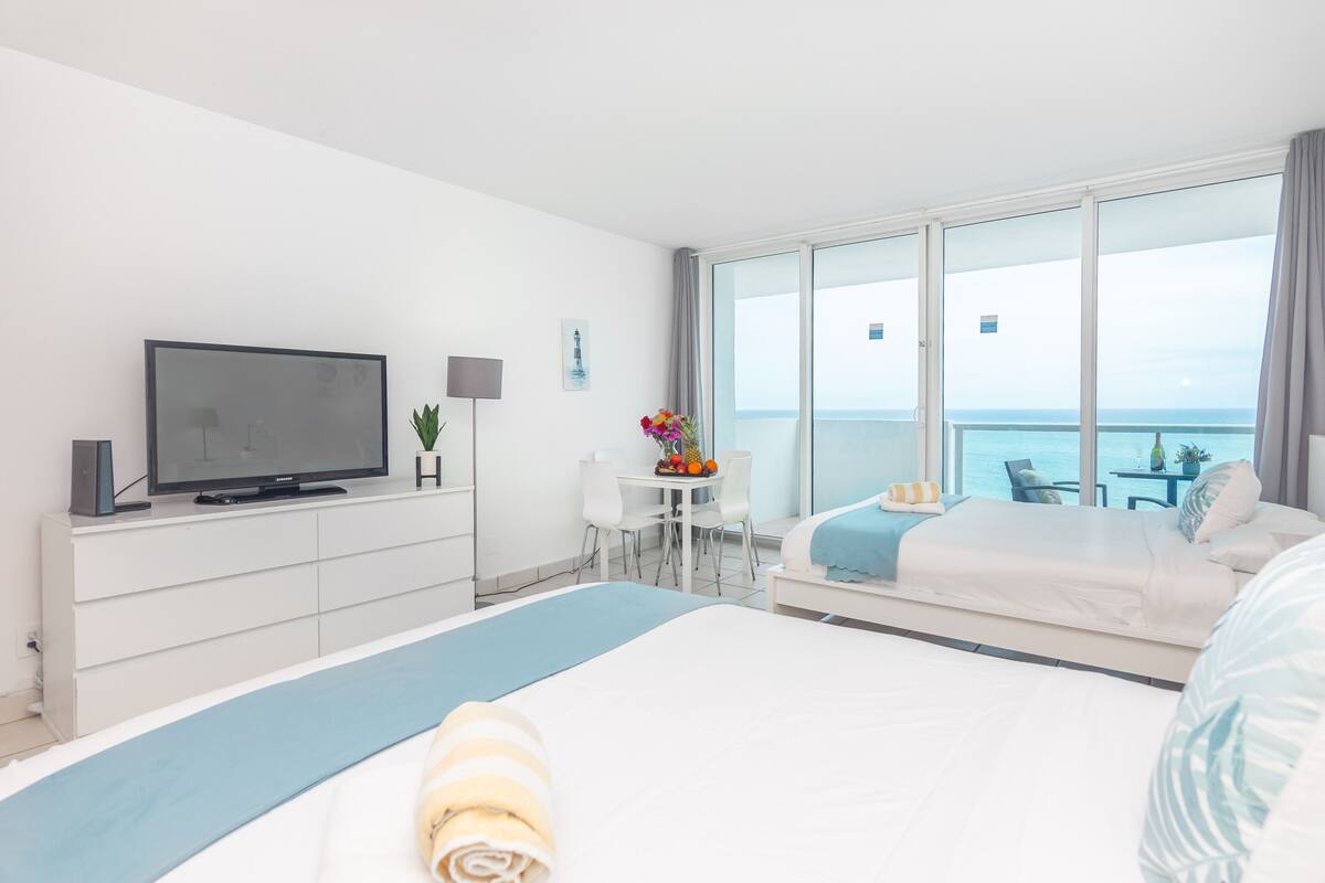 A hotel room with a large bed, a television, a small table with fruit, and a balcony with a view of the sea.