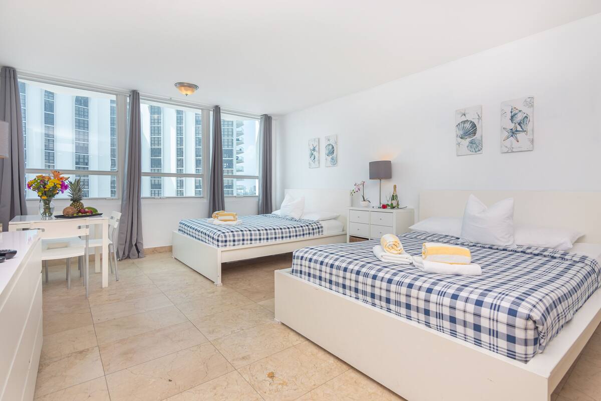 A modern hotel room with two beds, a desk, and a dining area.