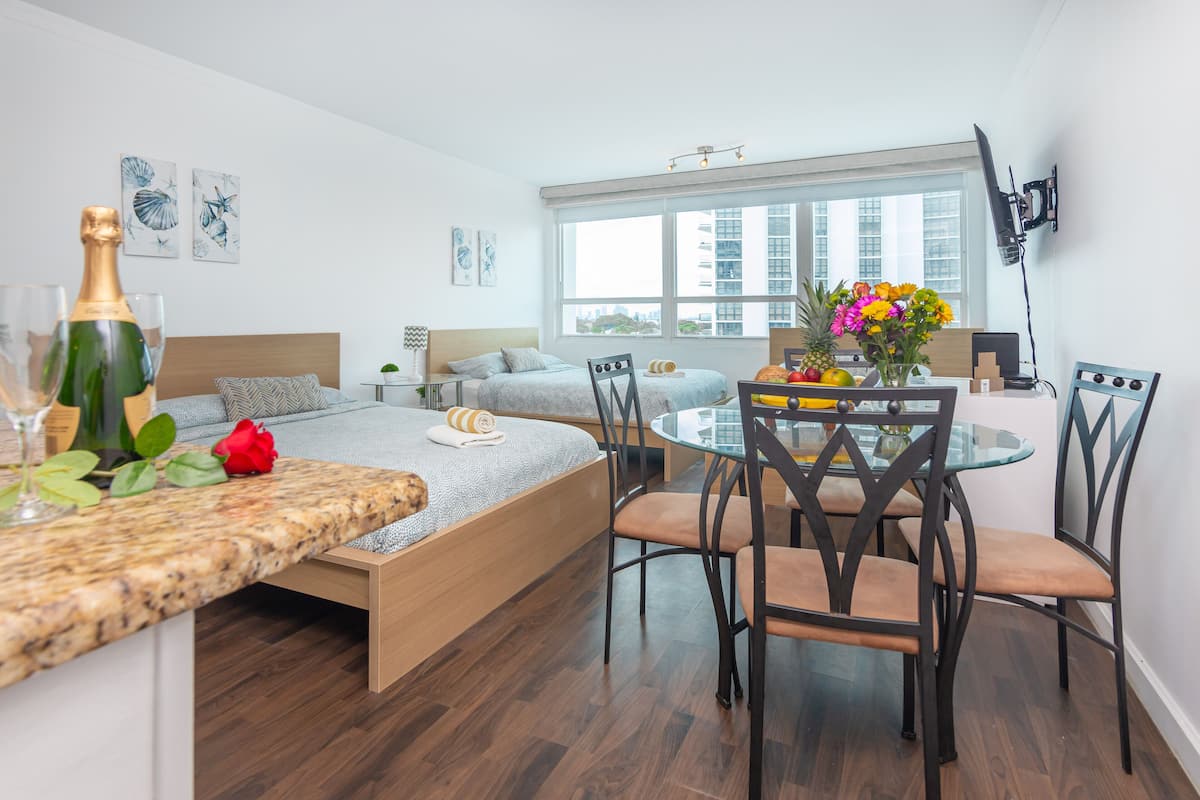 A modern hotel room with a bed, a dining table with chairs, a TV mounted on the wall, and artwork on the walls.