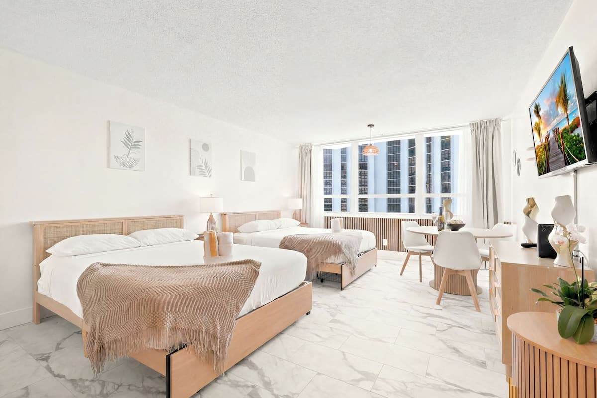 A hotel room with two beds, a desk, a chair, a TV, and a large window.