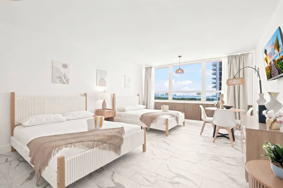 A modern hotel room with a large bed, a smaller bed, a desk, and a view of the city.