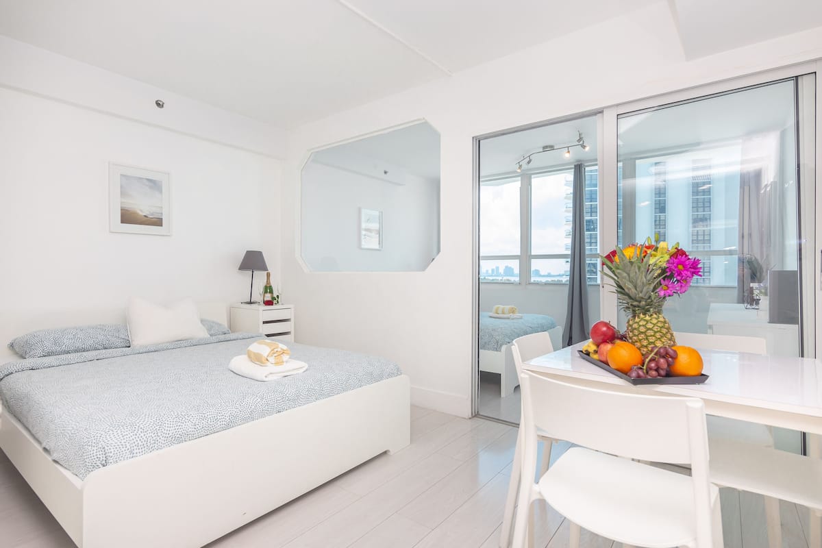 A modern bedroom with a bed, bedside table, and a dining area with a fruit bowl.