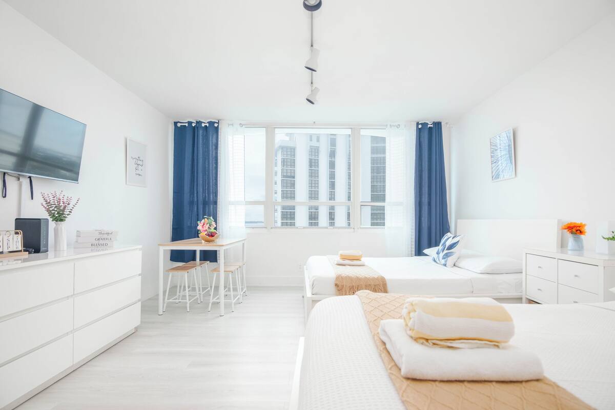 A modern hotel room with a large window, a desk with a chair, two beds, and a TV on the wall.