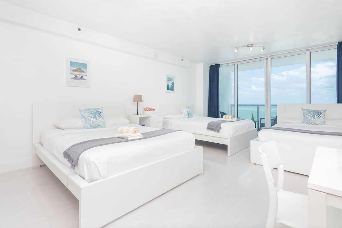 A modern hotel room with two beds, a desk, and large windows offering a view of the ocean.