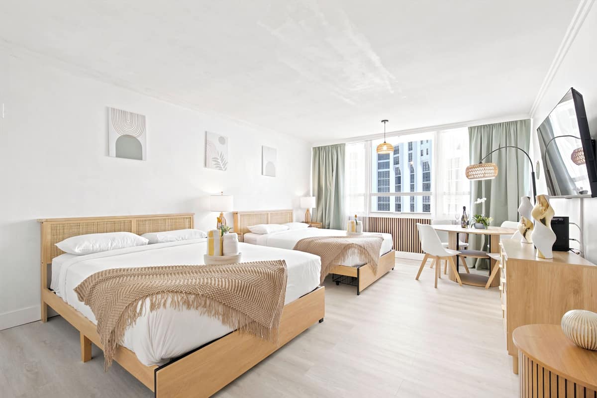 A hotel room with two beds, a desk, a chair, a TV, and a window with a view of buildings.