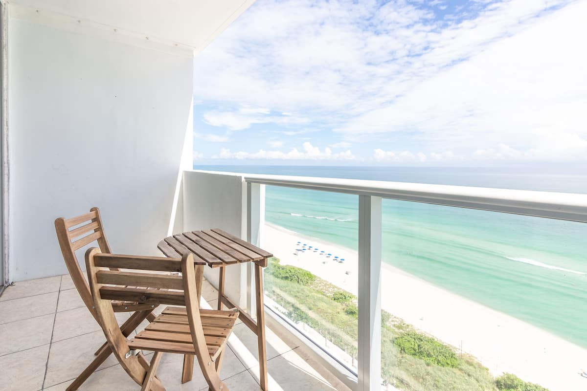 A balcony with a wooden table and chairs, overlooking a beach with clear turquoise water and a white sandy shore.
