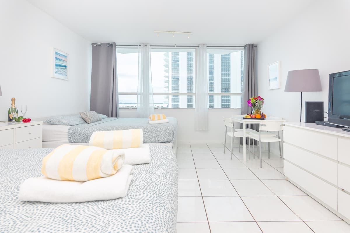 A modern hotel room with two beds, a dining area, a TV, and a large window with a city view.