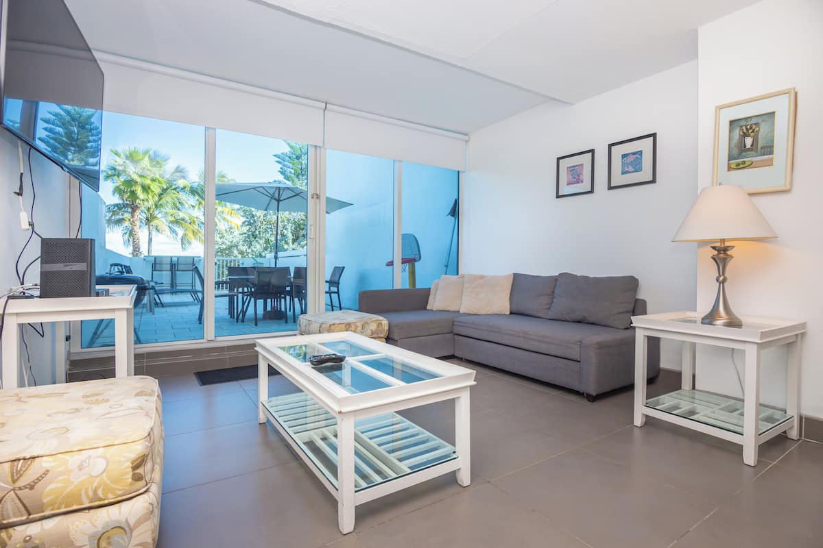 A modern living room with a glass coffee table, a sofa, and a side table with a lamp. There are framed pictures on the wall and a view of palm trees outside.