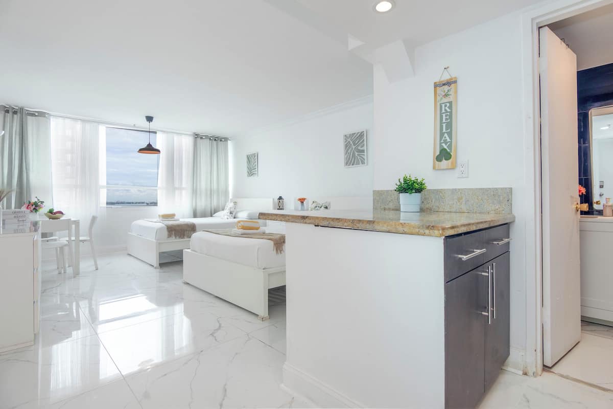 A modern, well-lit living area with a large window, a white sofa, a dining table, and a kitchen area with a countertop and cabinets.