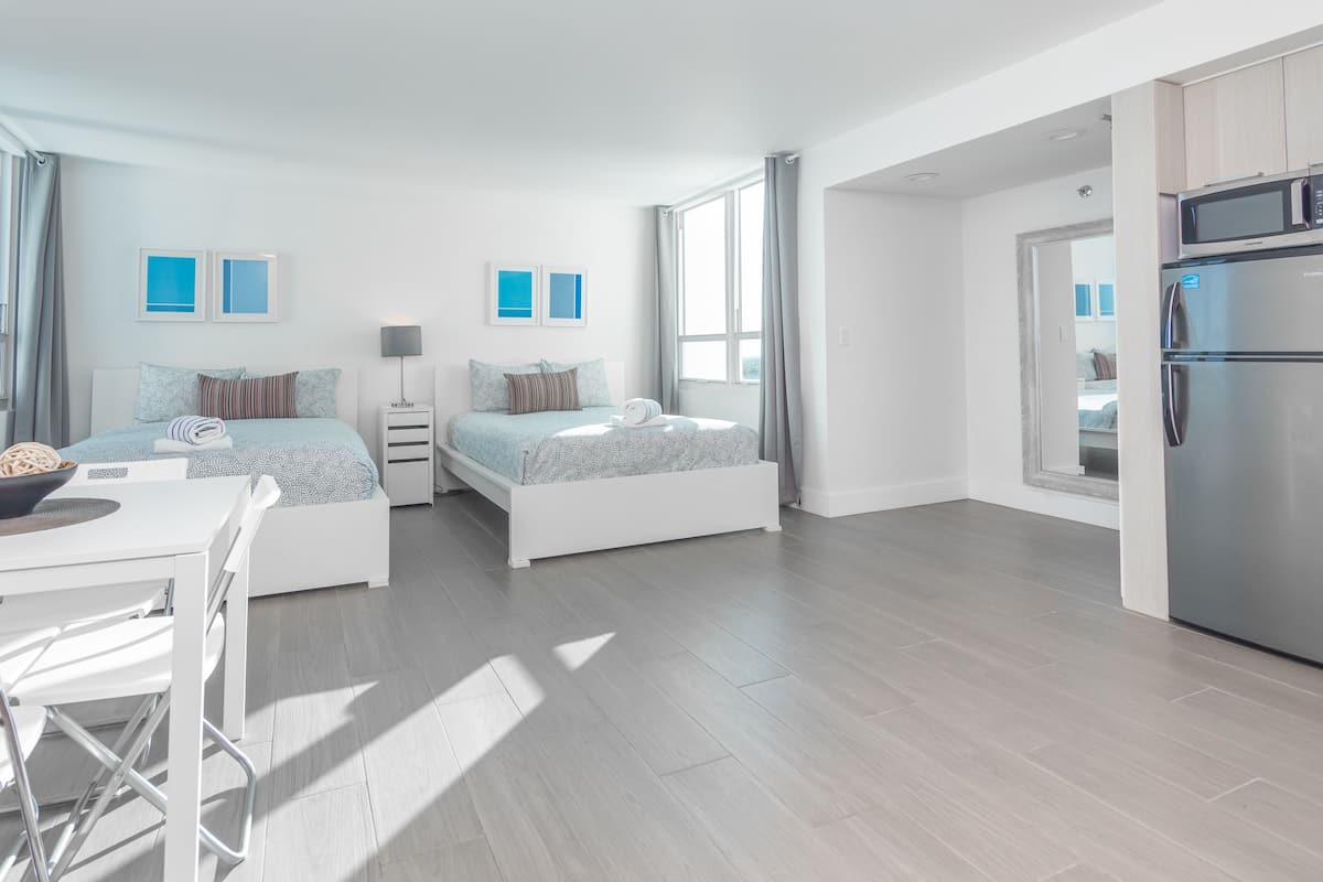 A modern hotel room with two beds, a dining area, and a kitchenette.