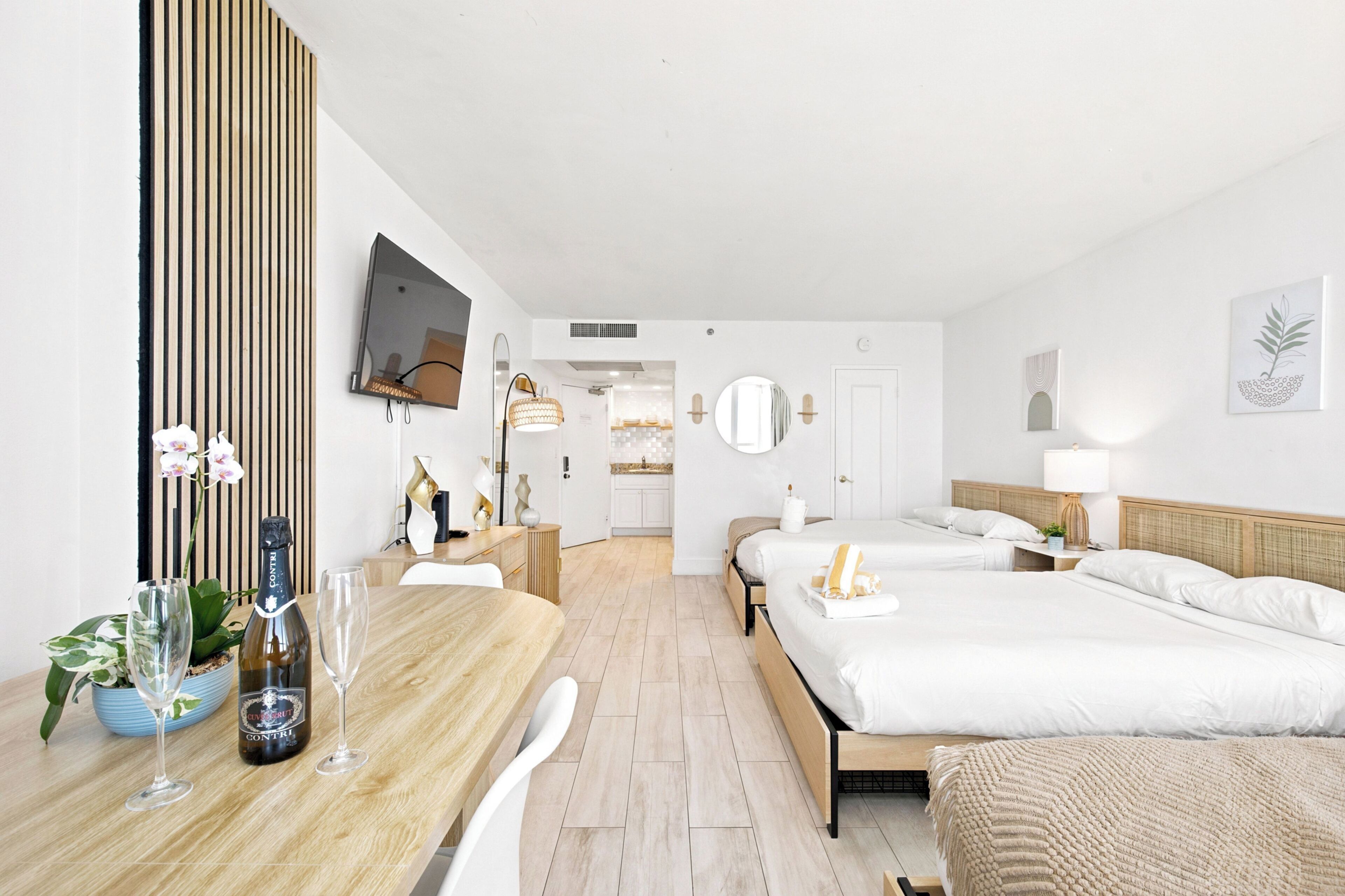 Una camera d'albergo con un tavolo da pranzo in legno, due letti, una TV e un bagno visibile attraverso una porta aperta.