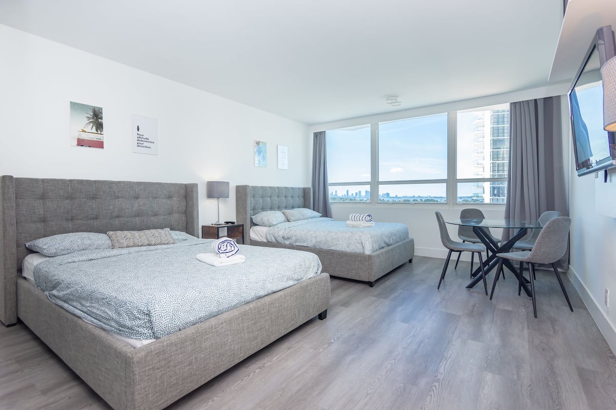 A modern hotel room with two beds, a dining area, and large windows offering a city view.