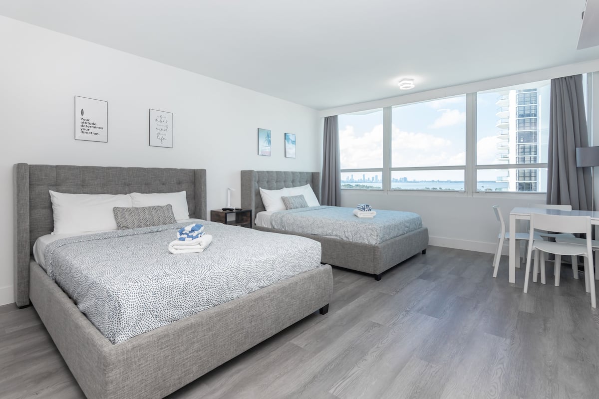 A modern hotel room with two beds, a dining area, and large windows offering a city view.