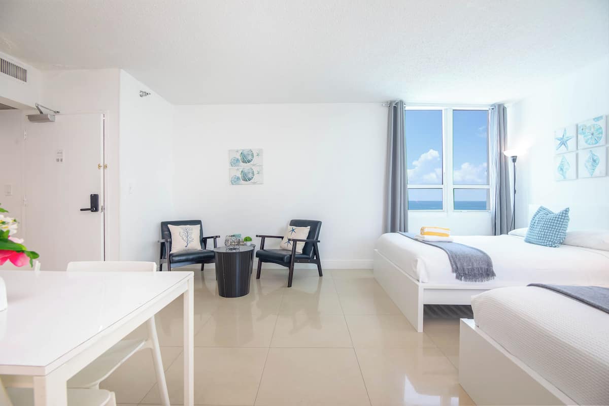 A modern hotel room with a bed, a desk, two chairs, a small table, and a window with a view of the sea.