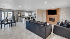 A modern living room with a fireplace, a dining area with a table and chairs, and a kitchen in the background.