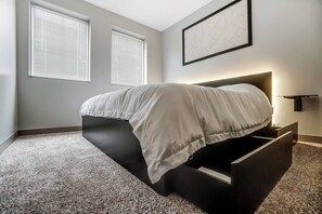 Un dormitorio moderno con una cama, dos ventanas, una obra de arte en la pared y una mesita de noche.