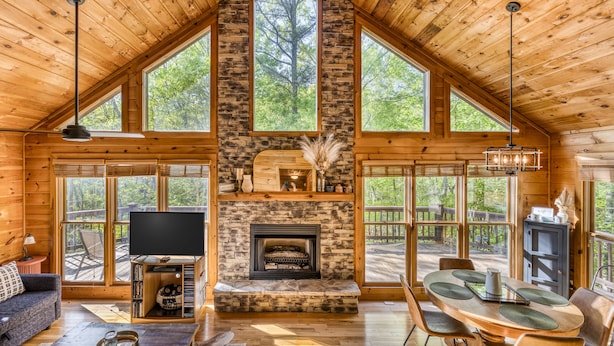 A log cabin with a stone fireplace, wooden floors, a dining area, and large windows offering views of the forest.